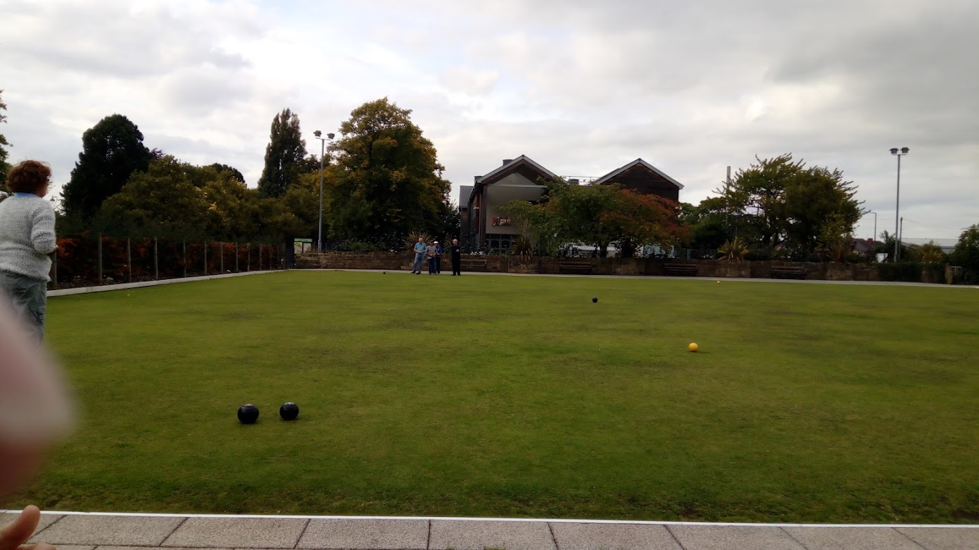 Green bowling green with players, trees, and buildings in the background under a cloudy sky.