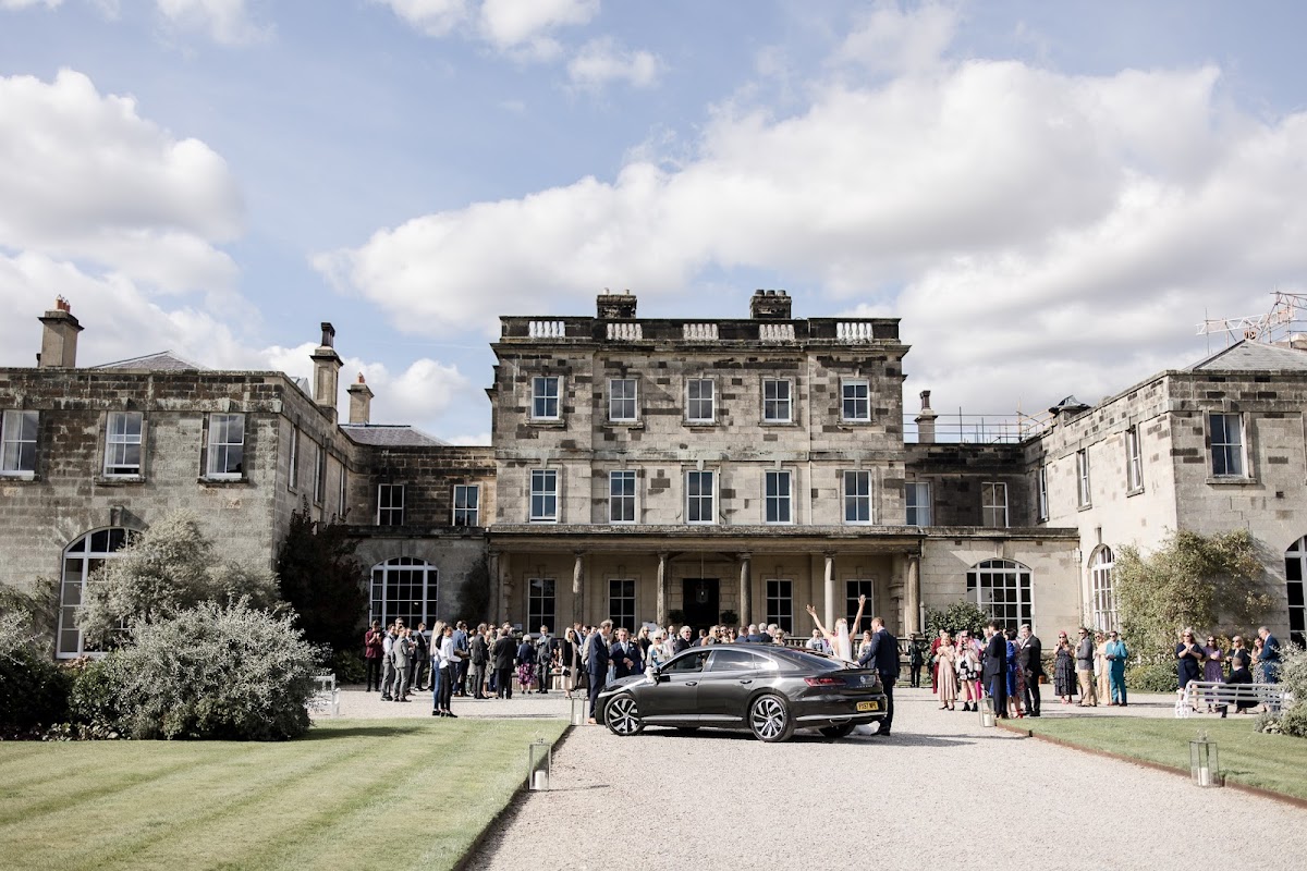 Elegant mansion with guests gathered outside, lush lawn, and a sleek car parked in front under a cloudy sky.