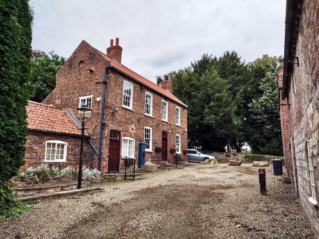 Charming brick hotel with red roof, surrounded by greenery and gravel driveway, featuring a lantern and parked car.