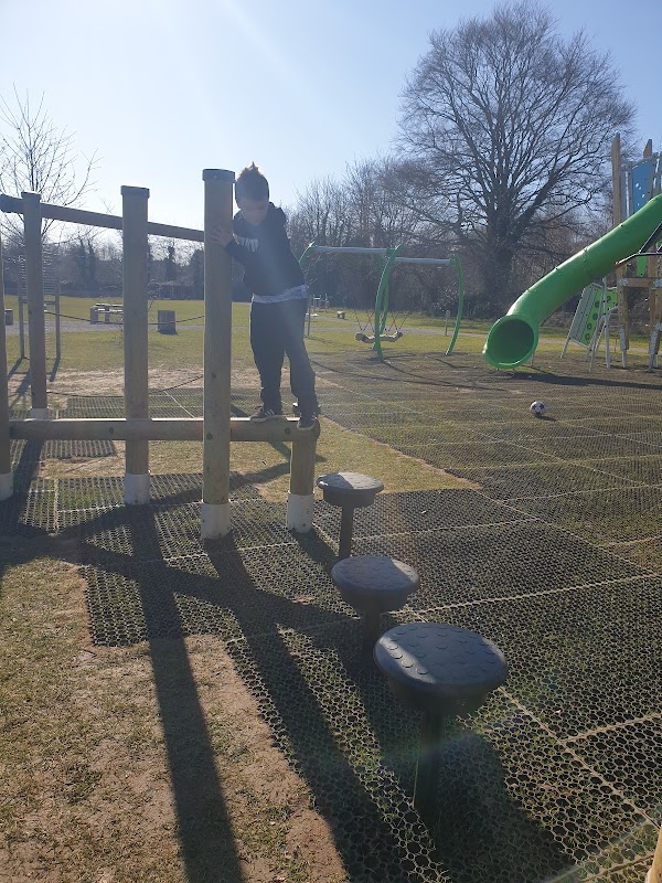 A child climbing on wooden playground equipment with a slide in the background, set in a grassy area under clear blue skies.