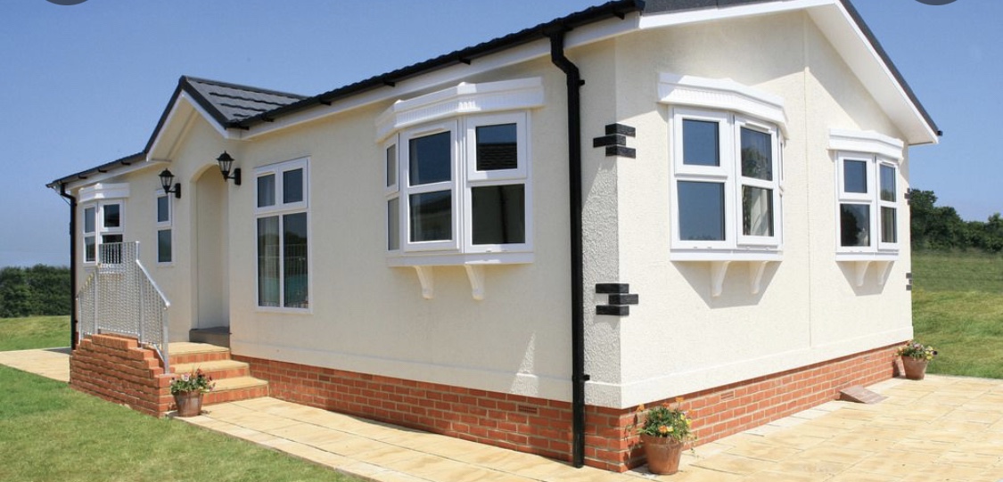 Modern mobile home with white exterior, multiple windows, and a brick patio surrounded by green grass.