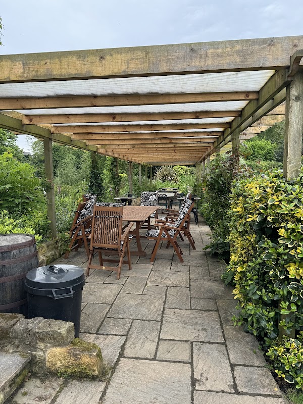 Outdoor dining area with wooden tables and chairs, surrounded by lush greenery and stone pathways.