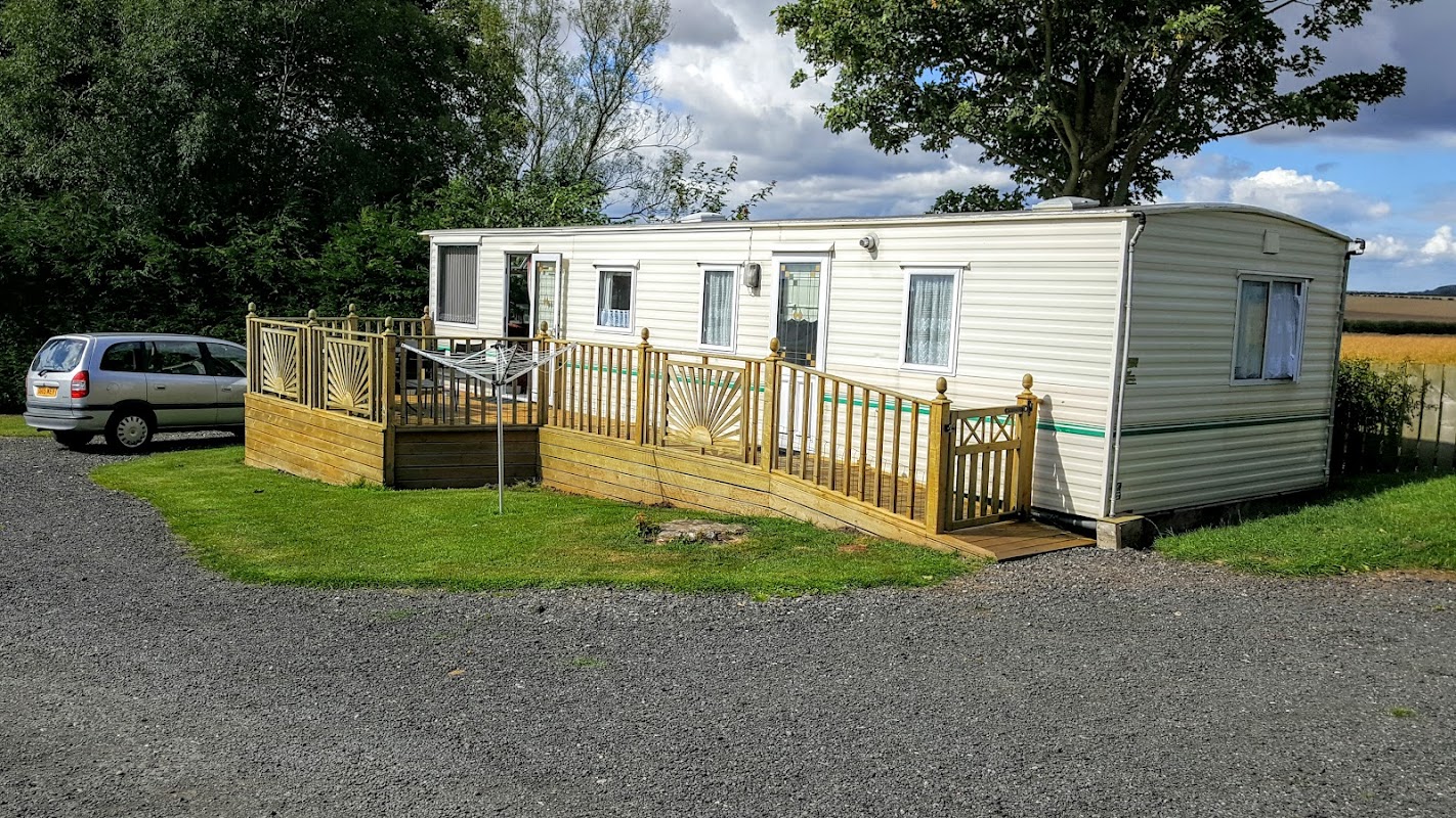 A mobile home with a wooden deck, surrounded by grass and trees, parked near a gravel road in a rural setting.