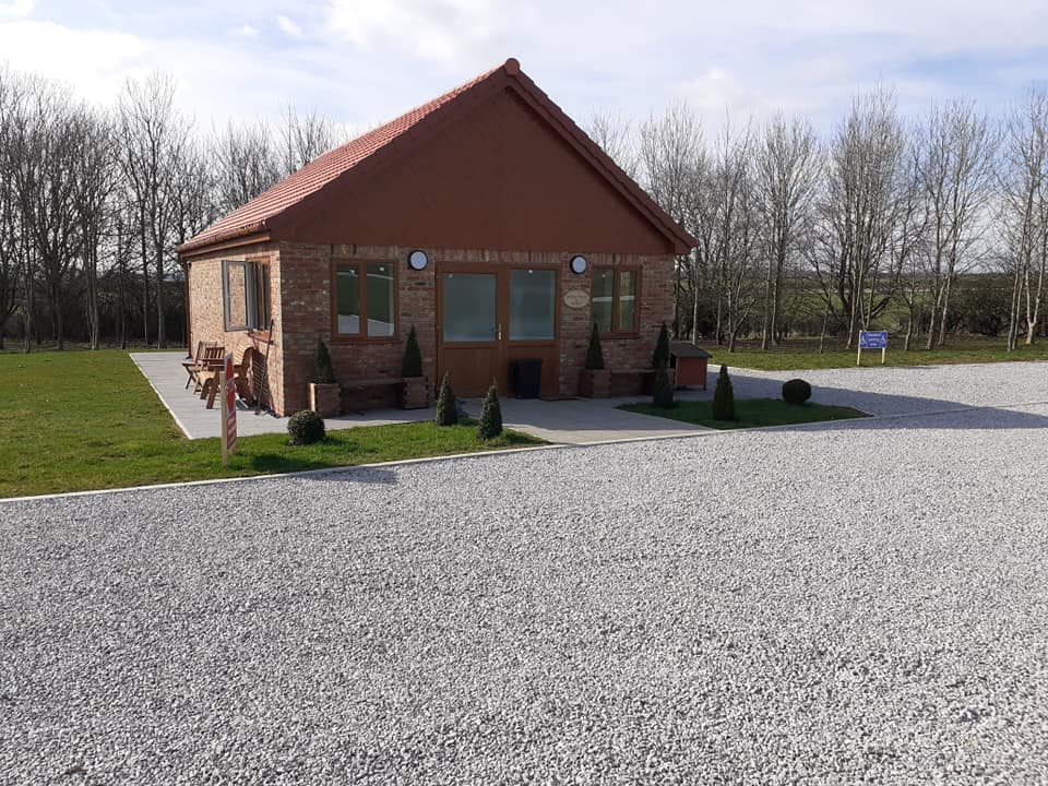 Quaint brick building with a red roof, surrounded by gravel and small shrubs, set against a backdrop of trees.