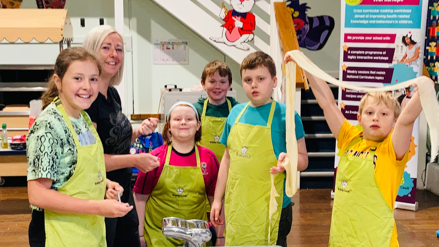Children in green aprons cooking with an instructor, preparing food in a bright, cheerful kitchen setting.