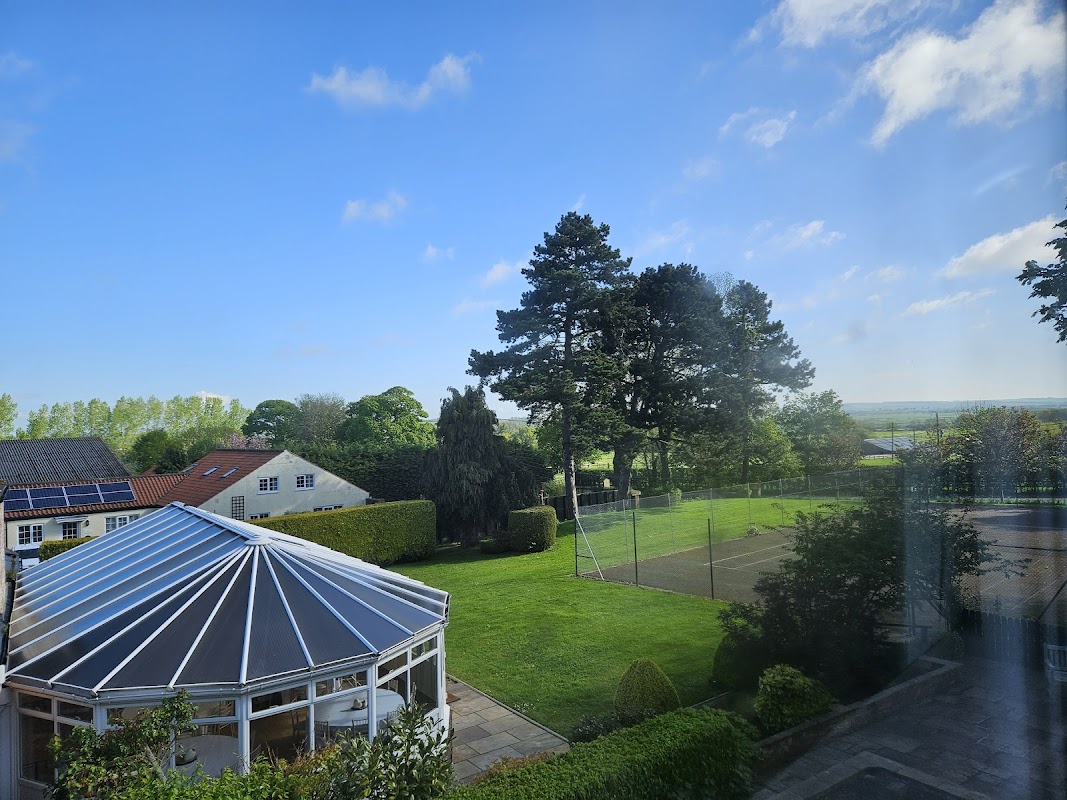 Burythorpe House features a conservatory, lush green lawn, tennis court, and trees under a clear blue sky.