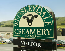 Wensleydale Creamery sign with cow logo against a backdrop of rolling green hills and blue sky.