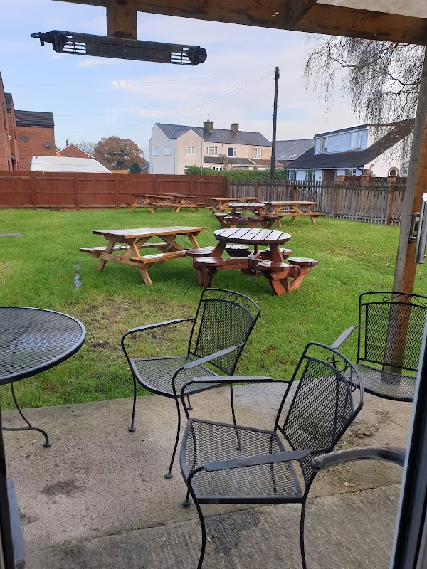 Outdoor seating area with picnic tables on green grass, surrounded by wooden fencing and residential buildings in the background.