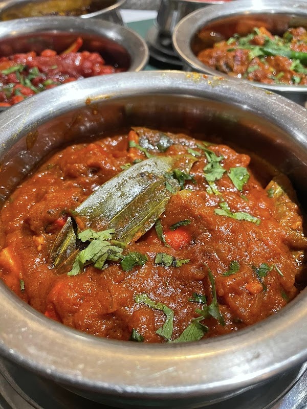 A rich, spicy curry topped with green herbs in a metal bowl, surrounded by other colorful dishes.