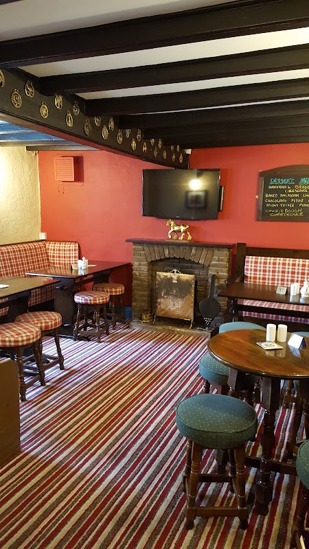 Cozy bar interior with striped carpet, wooden tables, plaid seating, a fireplace, and a TV on the wall.
