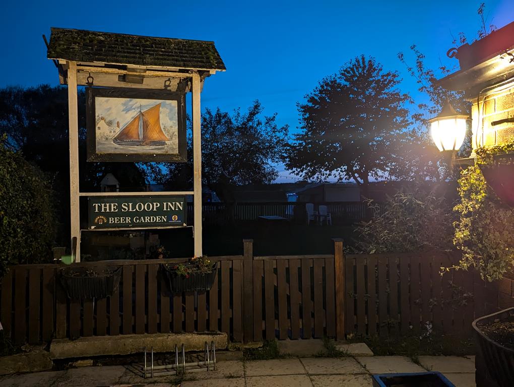 The Sloop Inn sign features a painting of a sailboat, surrounded by trees and a cozy beer garden at dusk.