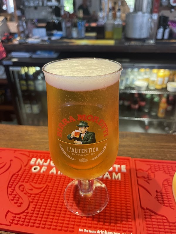 A pint of beer in a branded glass sits on a bar mat, with a blurred background of bottles and bar equipment.