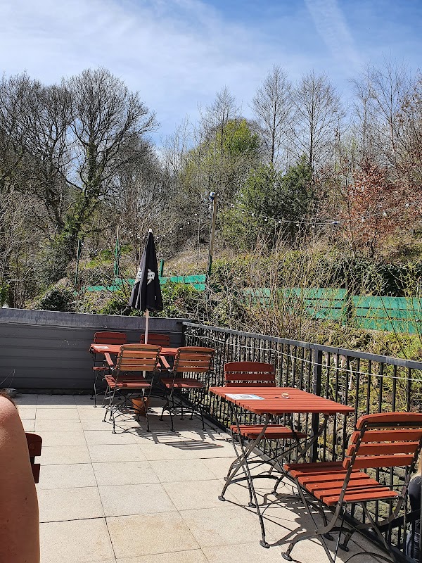 Outdoor seating area with wooden tables and chairs, surrounded by greenery and a black umbrella, under a clear blue sky.