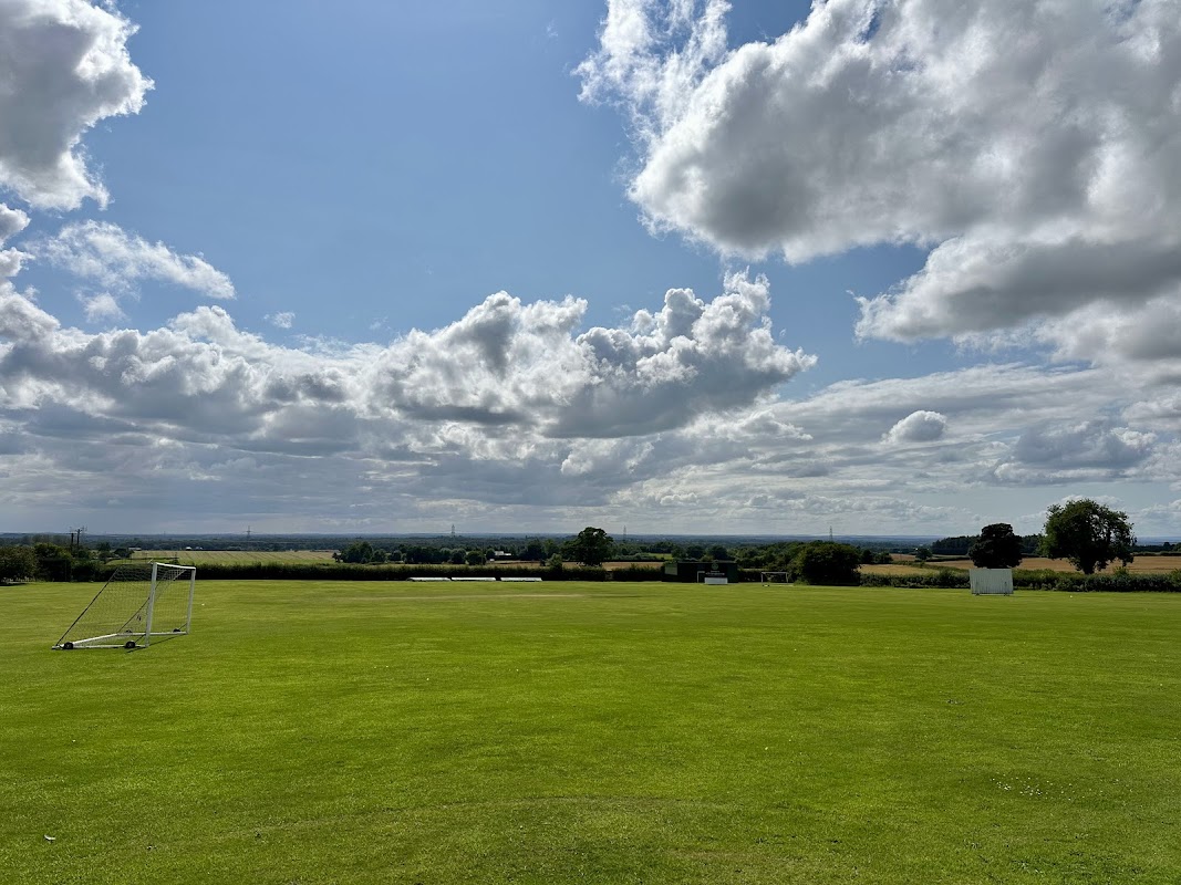 Green sports field with a goalpost, under a blue sky filled with clouds, overlooking a rural landscape.