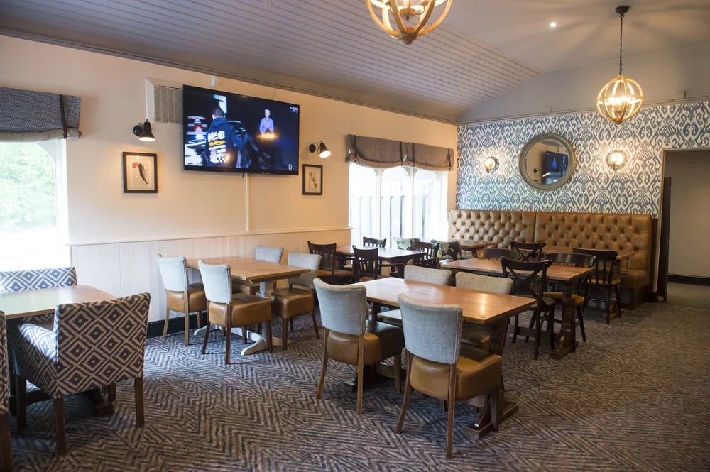 Cozy restaurant interior with wooden tables, chairs, patterned wallpaper, and a large TV on the wall.