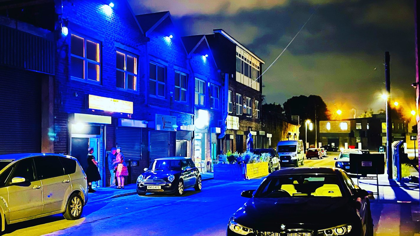 Nighttime scene of Tapestry Arts bar with blue lights, parked cars, and people outside in Dudley Hill, Yorkshire.