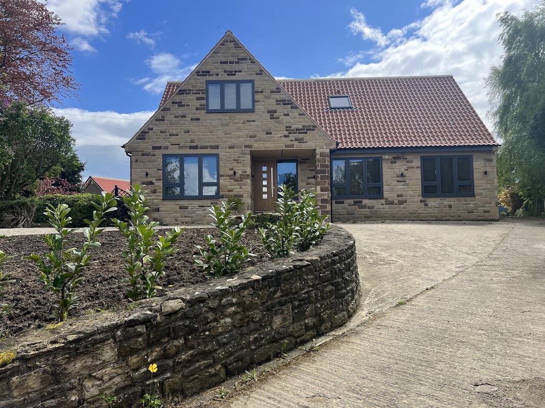 Modern stone house with a red tiled roof, landscaped garden, and a paved driveway under a blue sky.
