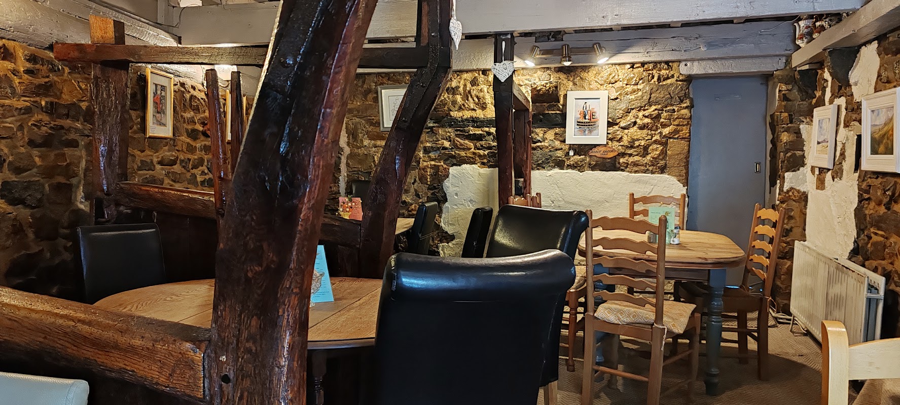 Cozy cafe interior with wooden beams, tables, chairs, and framed artwork on stone walls.