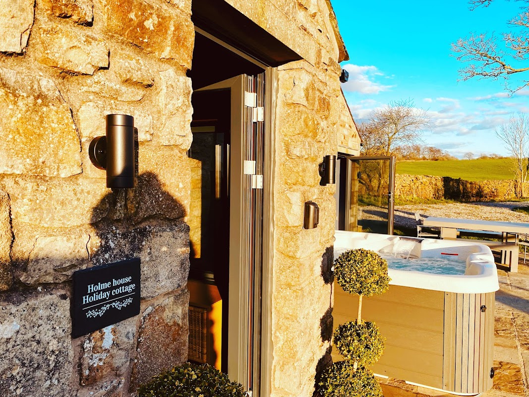 Stone cottage entrance with a sign, hot tub, and lush greenery under a bright blue sky.