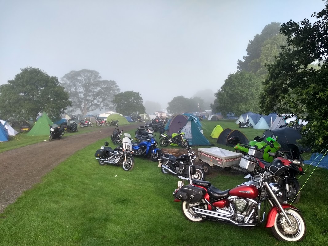 Tents and motorcycles scattered across a grassy field, surrounded by misty trees in a serene outdoor setting.