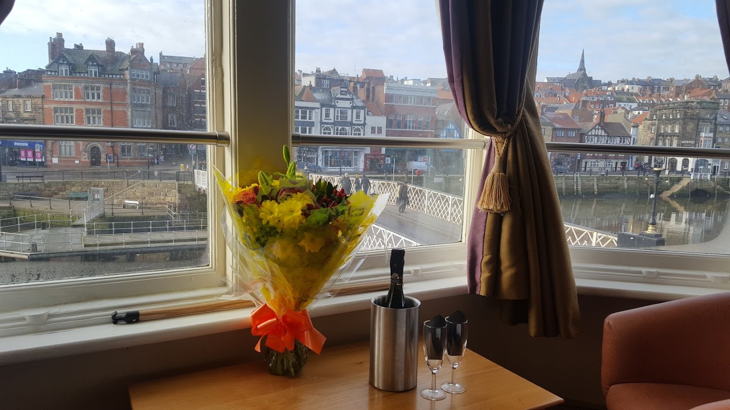 Bouquet of flowers and champagne glasses on a table by a window, overlooking Whitby’s colorful buildings and harbor.