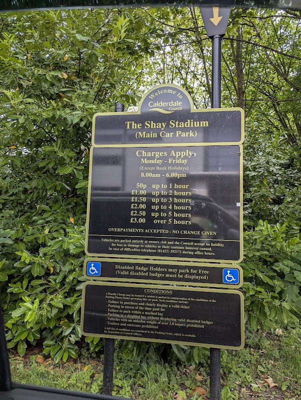 Sign detailing parking charges and conditions at Shay Stadium car park, surrounded by greenery in Exley, Yorkshire.