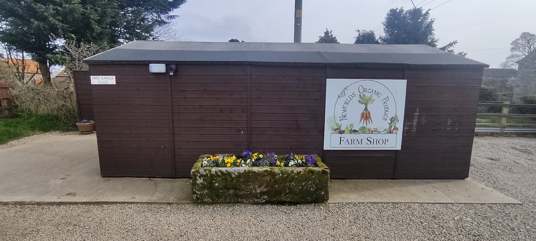 Wooden farm shop building with a sign reading "Newfields Organics" and a planter with colorful flowers at the entrance.