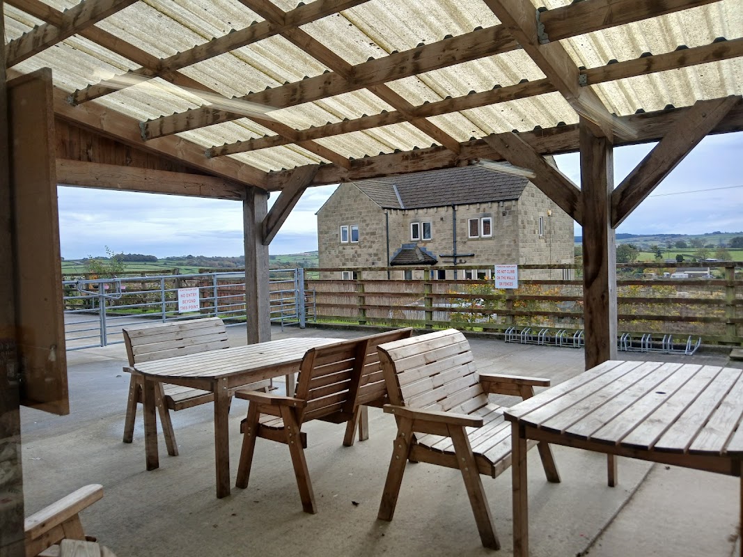 Wooden tables and benches under a covered patio, with a stone building and green hills in the background.