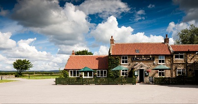 Charming stone inn with red-tiled roof, green umbrellas, and ivy, set against a vast, cloudy sky and open landscape.