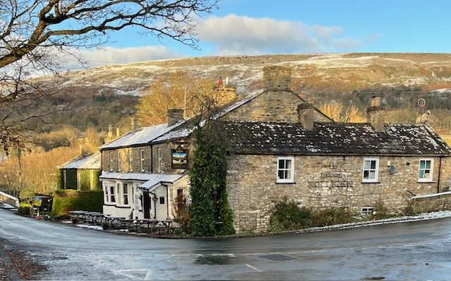Charming stone hotel with a sloped roof, surrounded by trees and hills, under a blue sky with scattered clouds.