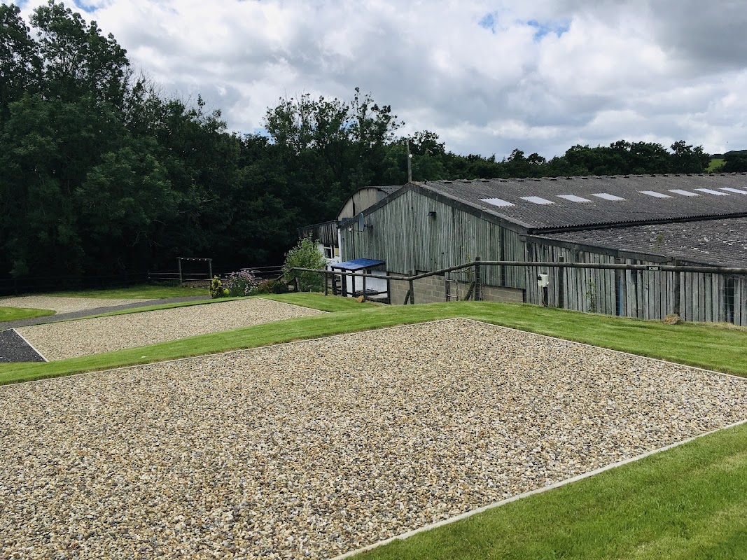 Gravel camping pitches with green grass, surrounded by trees and a rustic building under a cloudy sky.