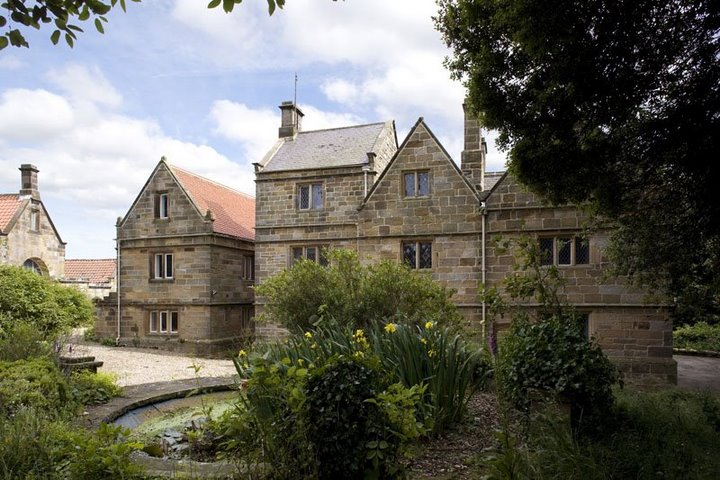 Historic stone building surrounded by lush greenery and a garden, with a blue sky and scattered clouds above.