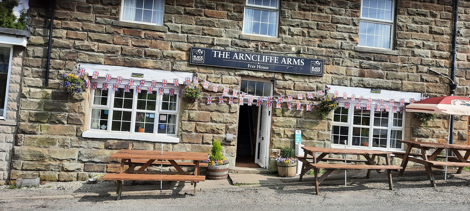 ARNCLIFFE ARMS B&B LTD sign, stone building, outdoor seating, flower pots, flags, and open door at 16:00.
