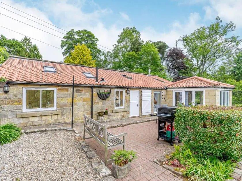 Charming stone cottage with a red-tiled roof, surrounded by greenery and a paved patio area with outdoor seating.