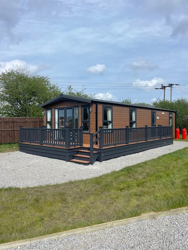 Modern holiday lodge with wooden exterior, deck, and gravel area, surrounded by greenery at The Meadows Holiday Park.