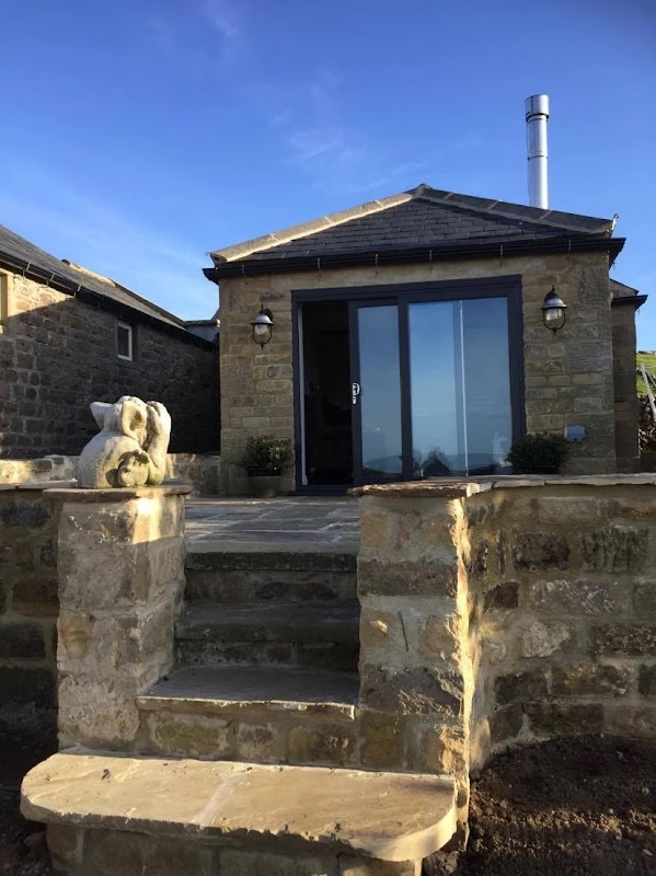 Stone cottage entrance with large glass doors, lanterns, and a stone staircase, set against a clear blue sky.