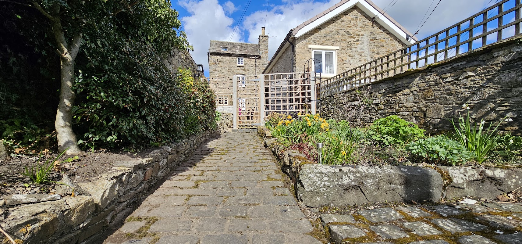 Pathway leading to Puzzle Cottage, flanked by stone walls and vibrant garden flowers under a blue sky.