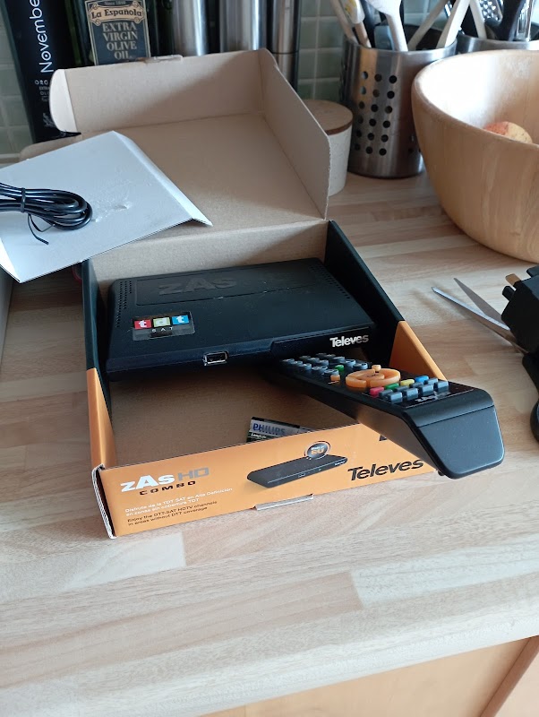 ZAS HD combo device in an open box with a remote control, on a wooden countertop with kitchen utensils in the background.