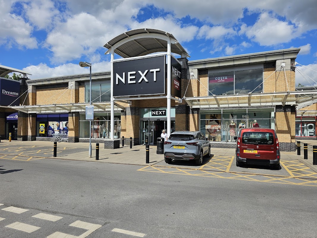 NEXT store at St James Retail Park, with parked cars and a blue sky in the background.