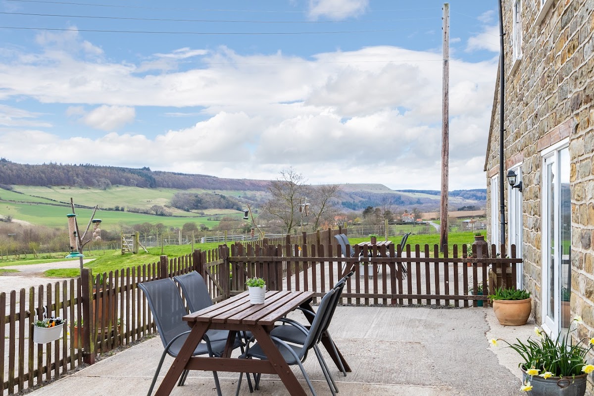 Outdoor seating area with wooden table and chairs, overlooking lush green hills and a cloudy sky in Harwood Dale.