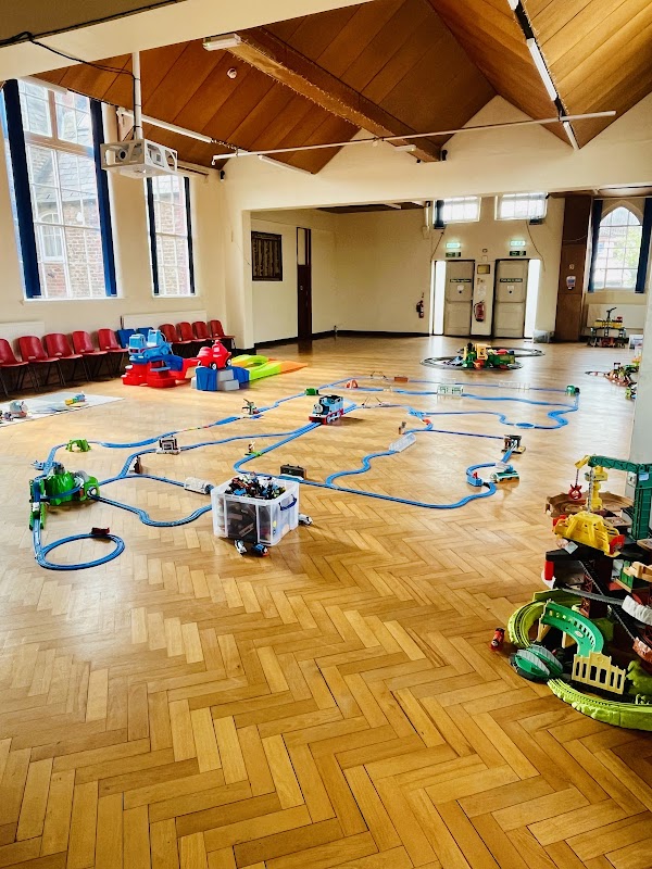 Bright hall with wooden flooring, toy train tracks, colorful playsets, and seating along the walls.