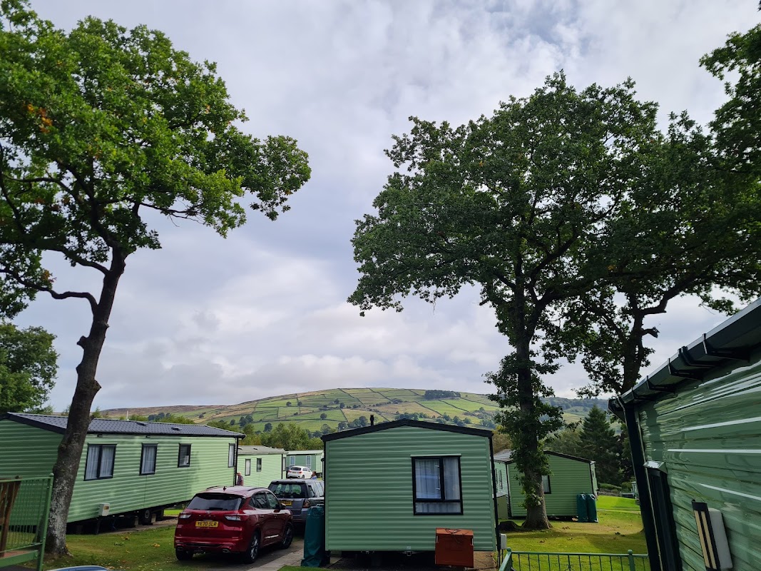 Green caravan units surrounded by trees, with rolling hills in the background under a cloudy sky.