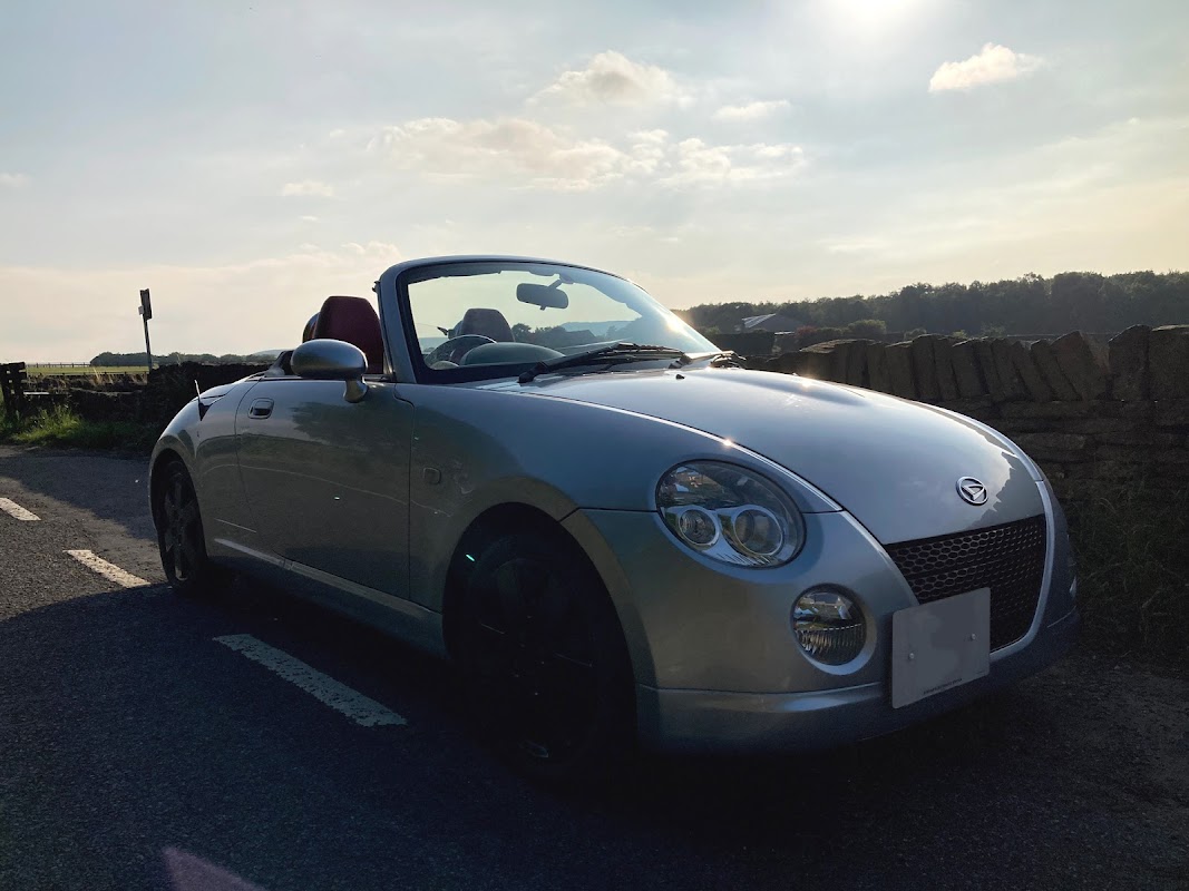 Silver convertible car parked on a country road with a scenic backdrop of hills and a blue sky.