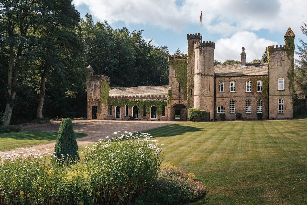 Victorian-style castle surrounded by lush greenery and manicured lawns, with a flag flying and flower beds in the foreground.