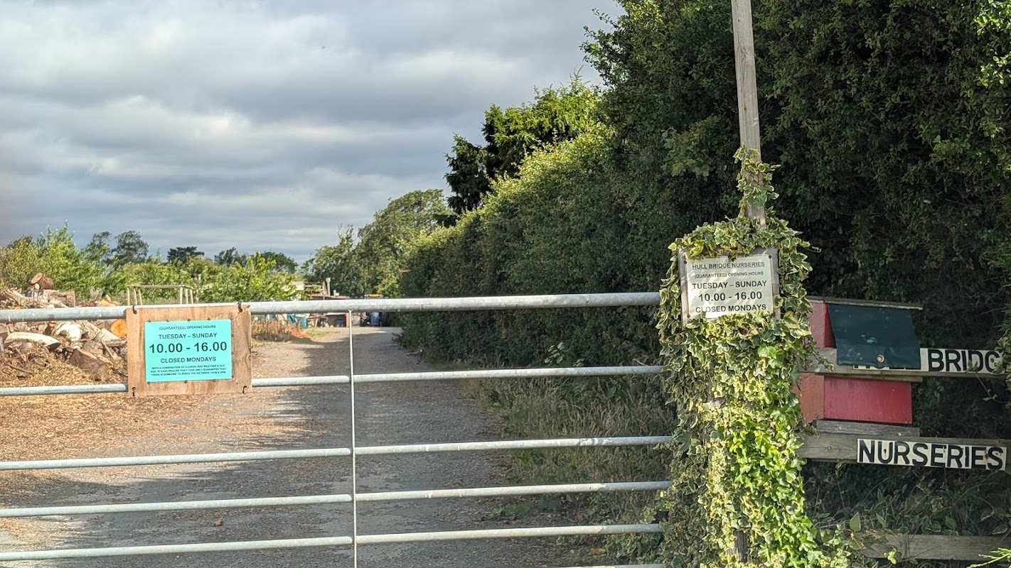 Gated entrance to Hullbridge Nurseries with opening hours signage and lush greenery along the pathway.