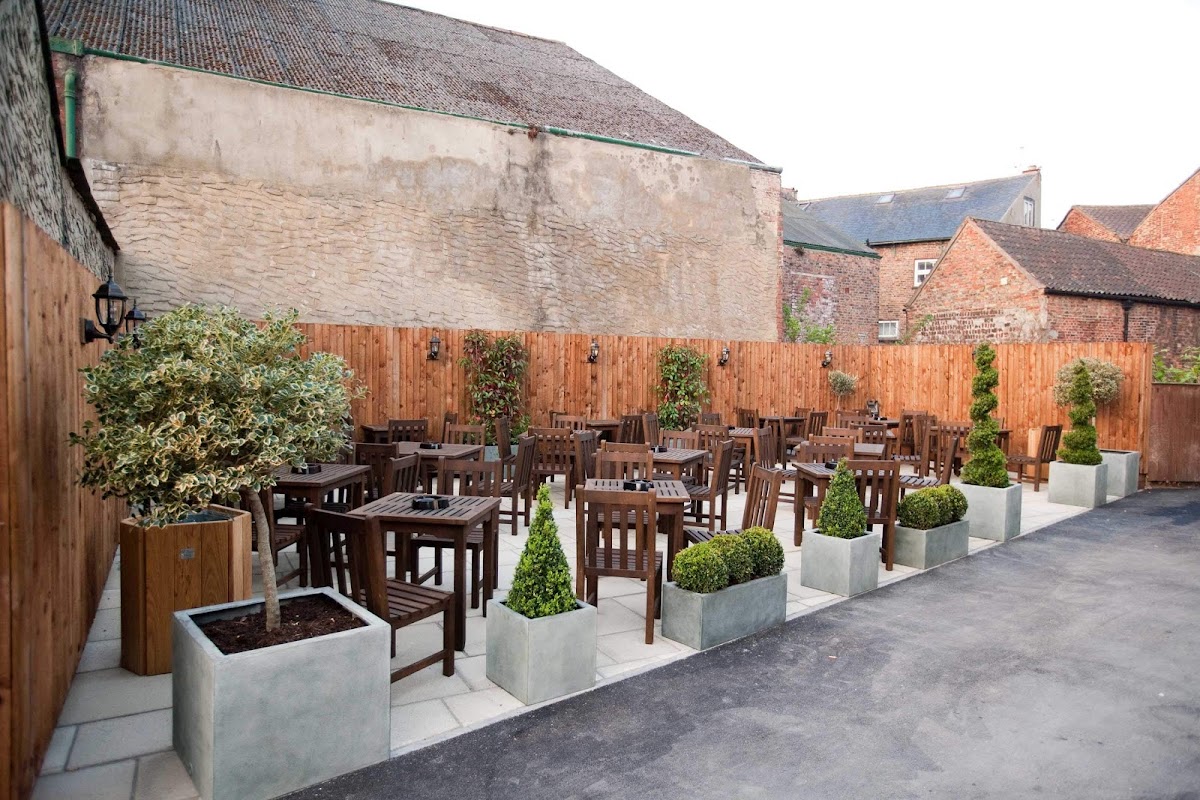 Outdoor dining area with wooden tables and chairs, surrounded by planters and greenery in a rustic setting.