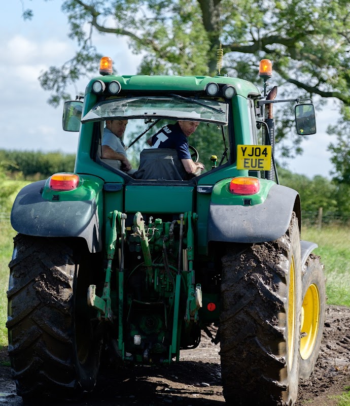 Two men in a green tractor on a dirt path, surrounded by lush greenery and blue skies in Yorkshire.