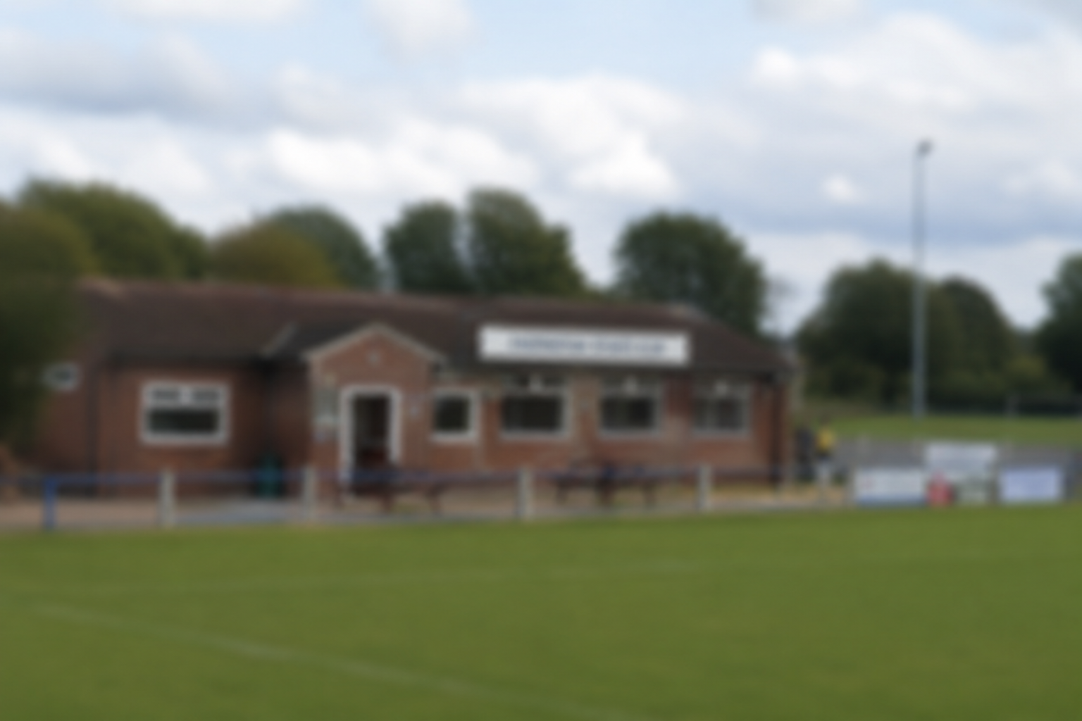 Modern cricket club building with a sign, outdoor seating, and people enjoying the space on a grassy field.