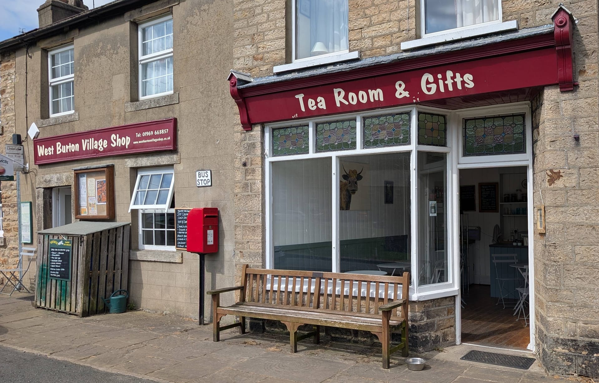 West Burton Village Shop and Tea Room with a bus stop, wooden bench, and parked car outside a stone building.
