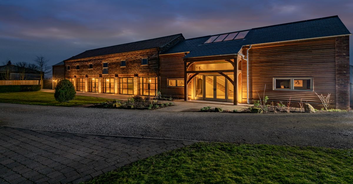Rustic event venue with wooden exterior, large glass doors, and landscaped garden, illuminated at dusk in Kelfield.